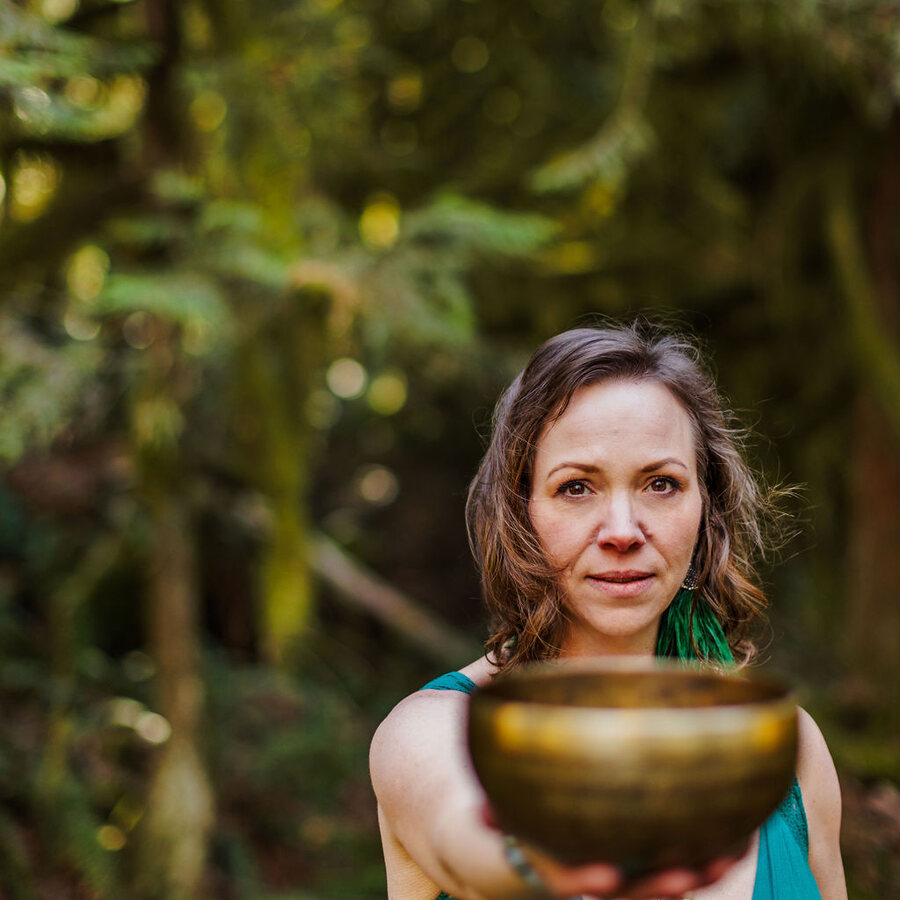 Angela Bolton in teal holding a bronze singing bowl out toward the camera in a green forest