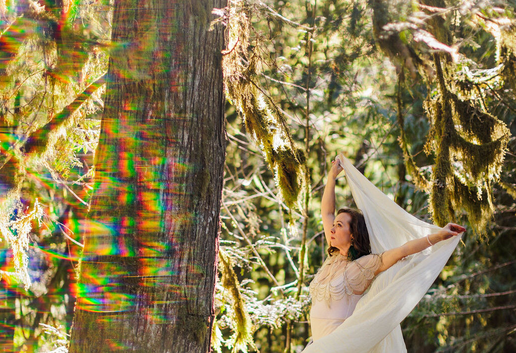 Rainbow light refracting across a moss-covered cedar, Angela Bolton in ivory beyond it