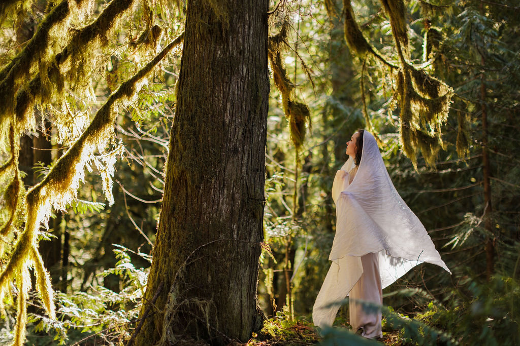 Angela Bolton in a mossy old-growth forest, a white veil lifting from her shoulders in low sun