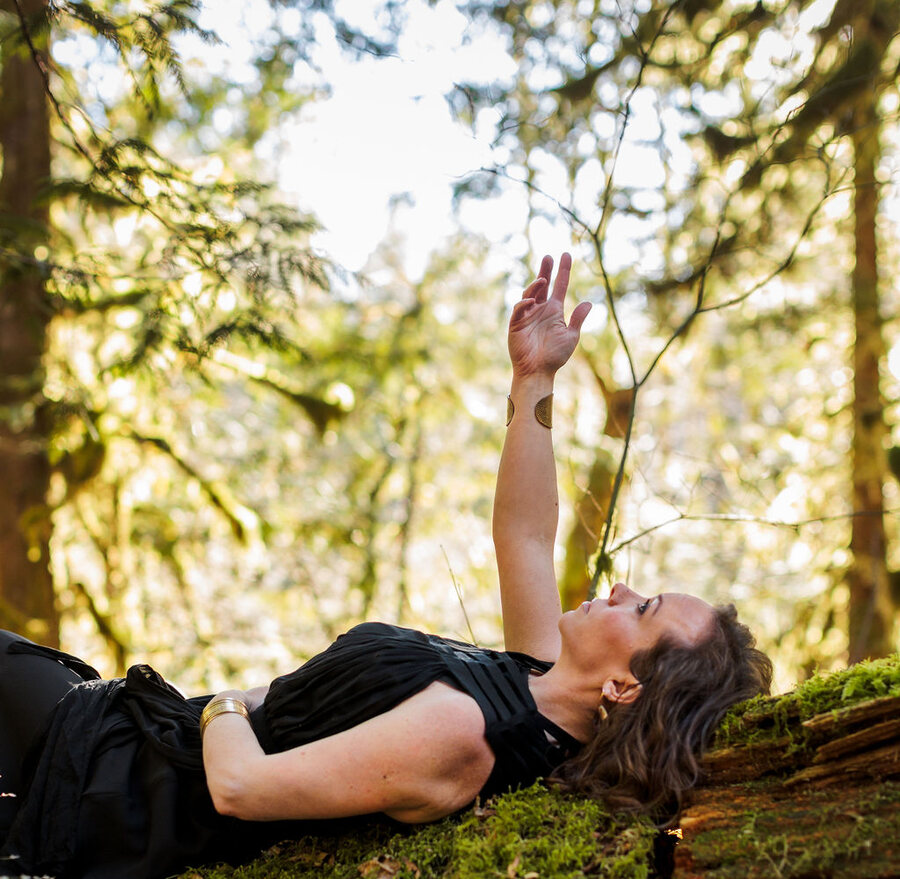 Angela Bolton lying back along a moss-covered log in a black dress, one arm reaching up into the light