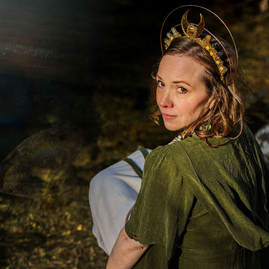 Angela Bolton seated at the water in a crescent-moon crown, looking back over her shoulder