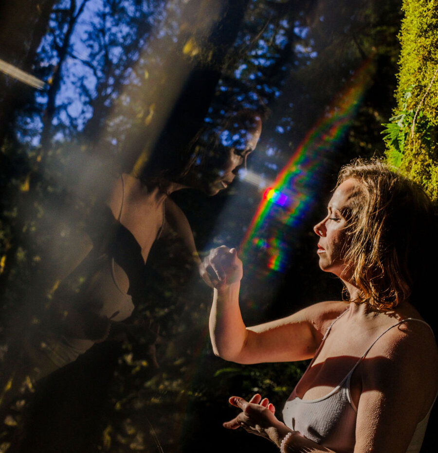 Angela Bolton and her own mirrored reflection facing each other through prismatic light in a dark forest