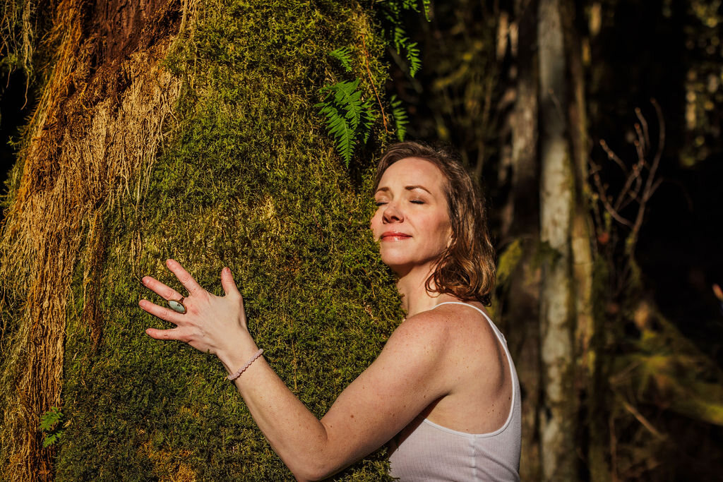 Angela resting her hands and cheek against a moss-covered trunk, eyes closed in the sun