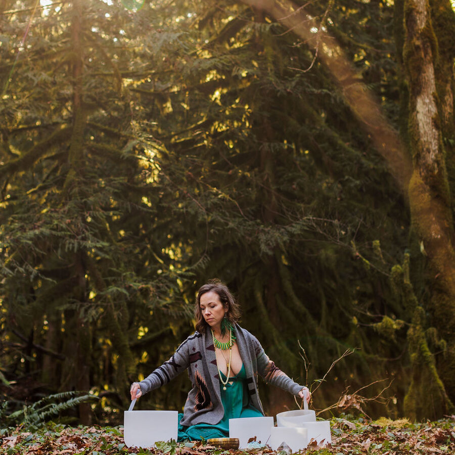 Angela Bolton seated on the forest floor playing two crystal singing bowls in low sun