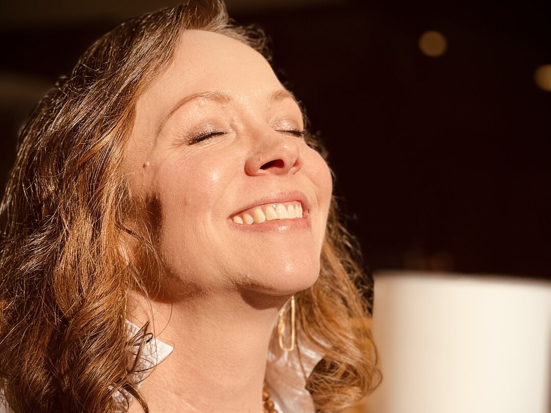 Angela Bolton in a close indoor portrait, eyes closed and smiling, warm lamplight behind her