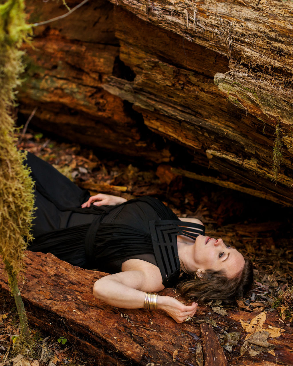 Angela lying back along a fallen cedar in a black dress, held by the forest floor